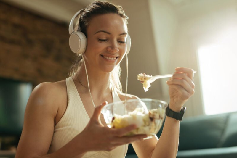 happy-athletic-woman-eating-salad-while-listening-music-headphones-relaxing-home